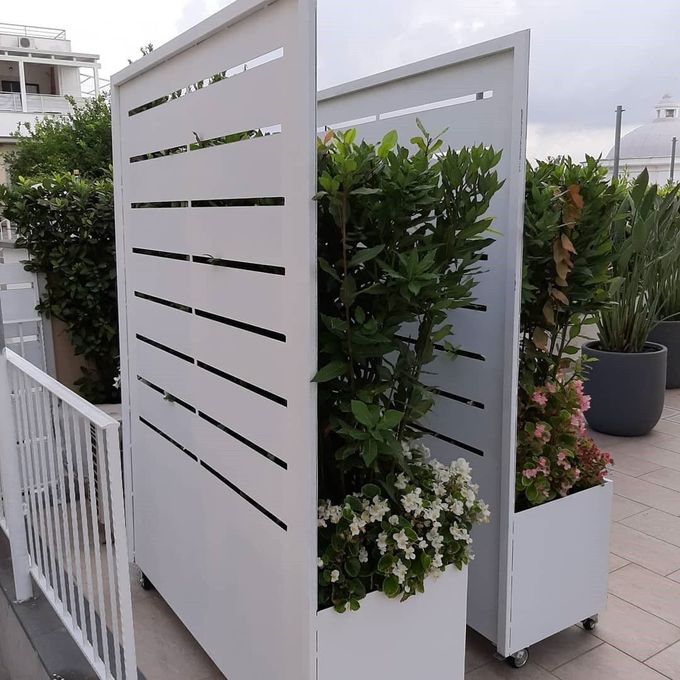 Su un balcone, due schermi frangivista in metallo bianco con fioriere integrate, riempiti di arbusti verdi e fiori bianchi.