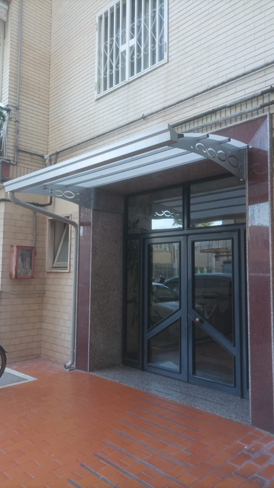 Una moderna pensilina in vetro e metallo sopra l'ingresso di un edificio con doppie porte in vetro grigio scuro e una rampa piastrellata di rosso.