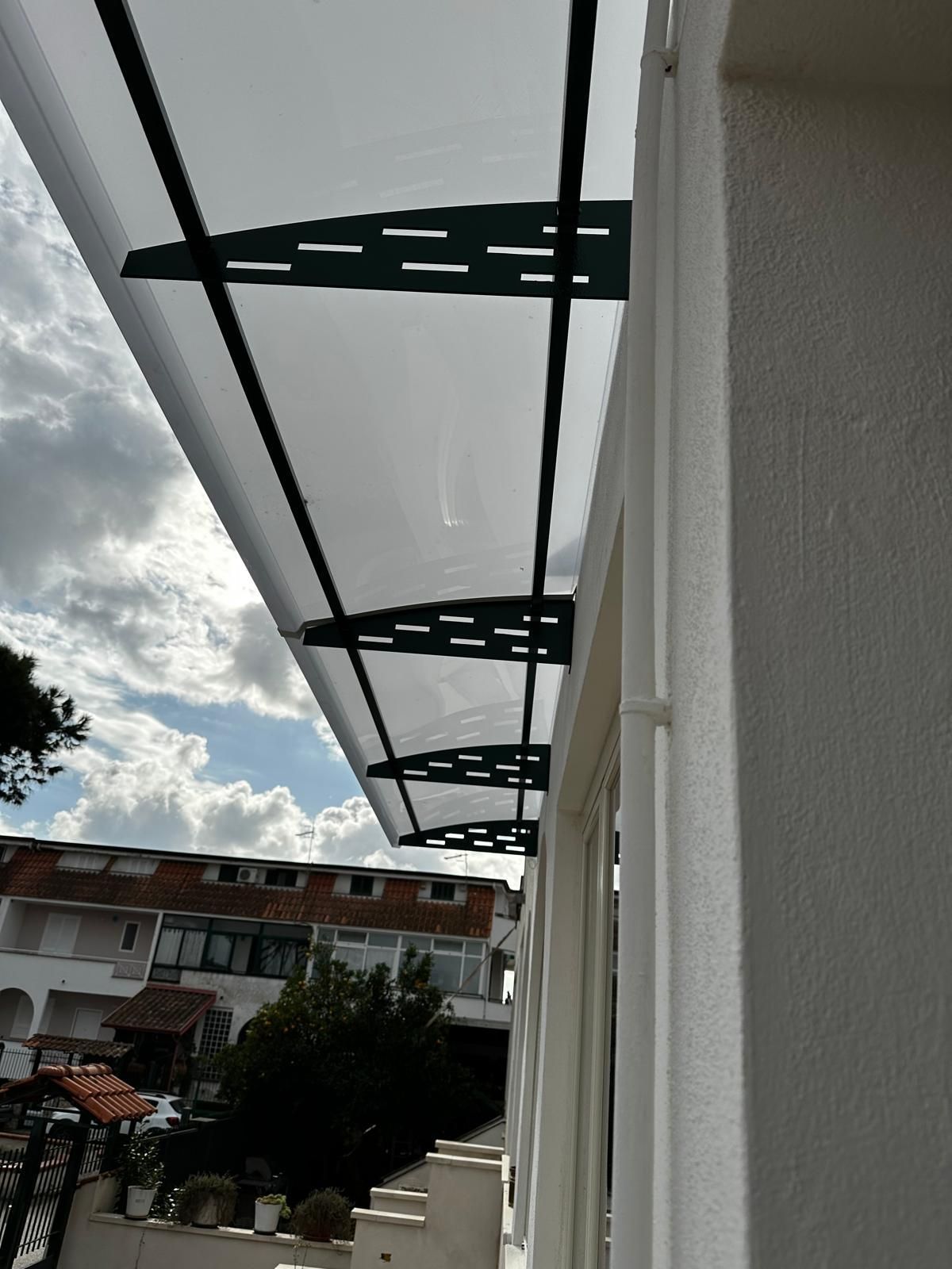 Una tettoia bianca e traslucida si estende da una parete dalla superficie ruvida, sorretta da staffe triangolari decorative in metallo scuro.