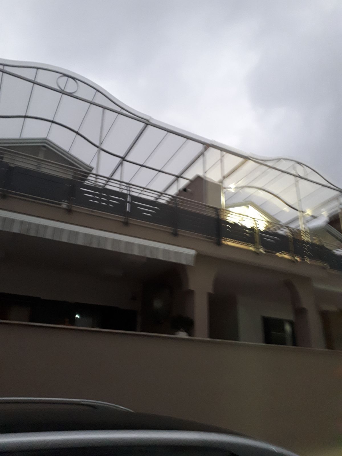 Un edificio a due piani color beige con una struttura del tetto in metallo bianco sopra un balcone esterno, visto dalla strada.