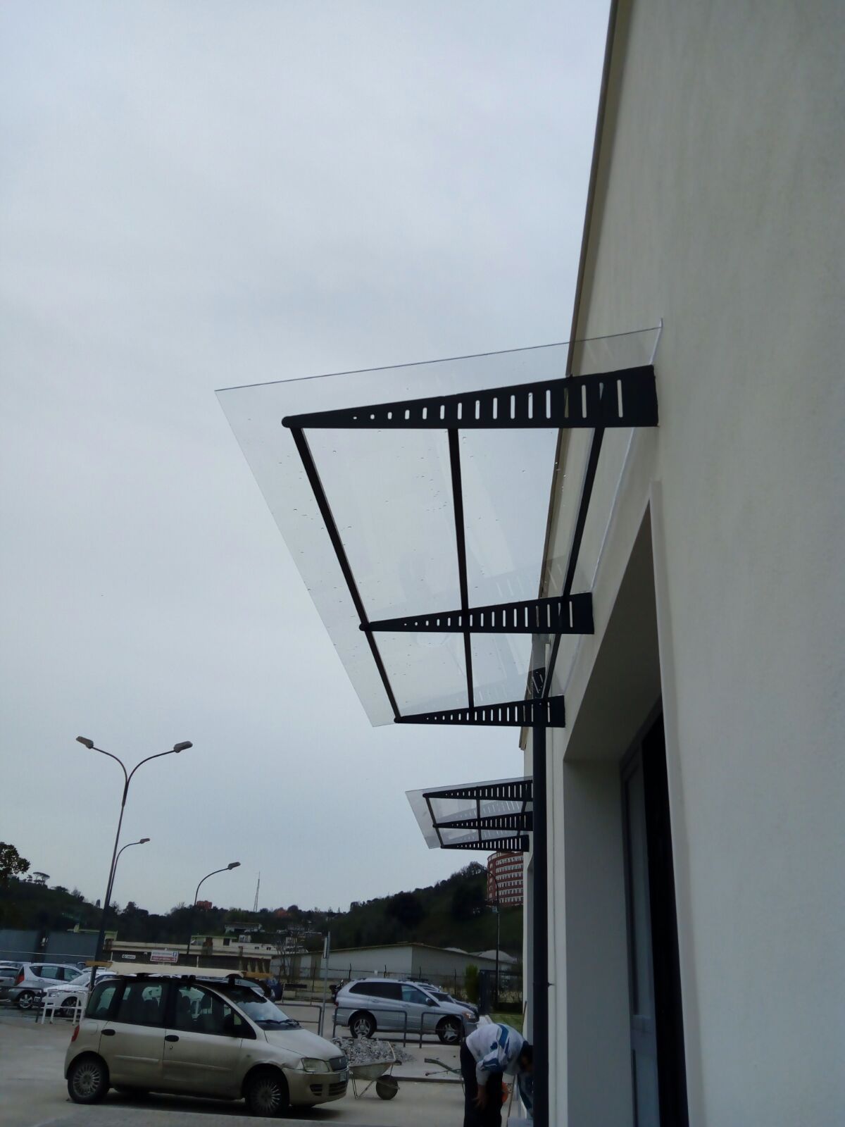 Una tettoia in metallo nero con una copertura in vetro trasparente fissata alla parete esterna di un edificio bianco in un parcheggio.