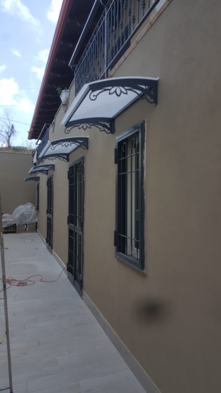 Una fila di finestre e una porta su una parete esterna beige, ciascuna sormontata da una tenda da sole in policarbonato con telaio nero abbinato.