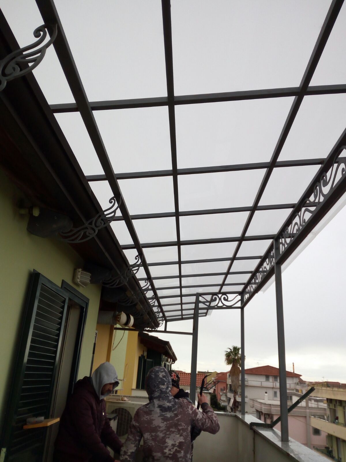 Due persone sono in piedi su un balcone sotto una tettoia con struttura metallica e tetto traslucido, installata di recente.