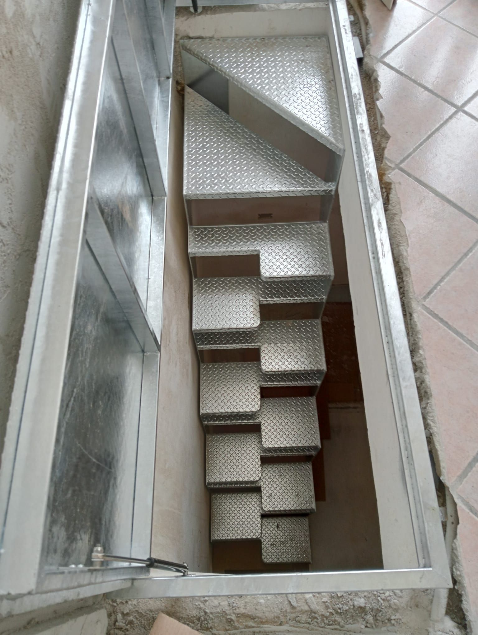 Una ripida e stretta scala in metallo argentato a piastra diamantata conduce, attraverso una botola sul pavimento, in un oscuro scantinato.