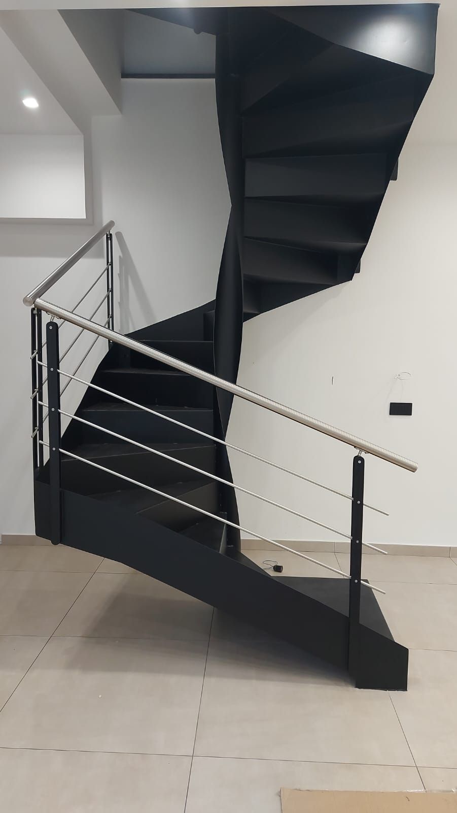 Una moderna scala a chiocciola nera con ringhiere in cavi metallici, in contrasto con pareti bianche e pavimento piastrellato chiaro.