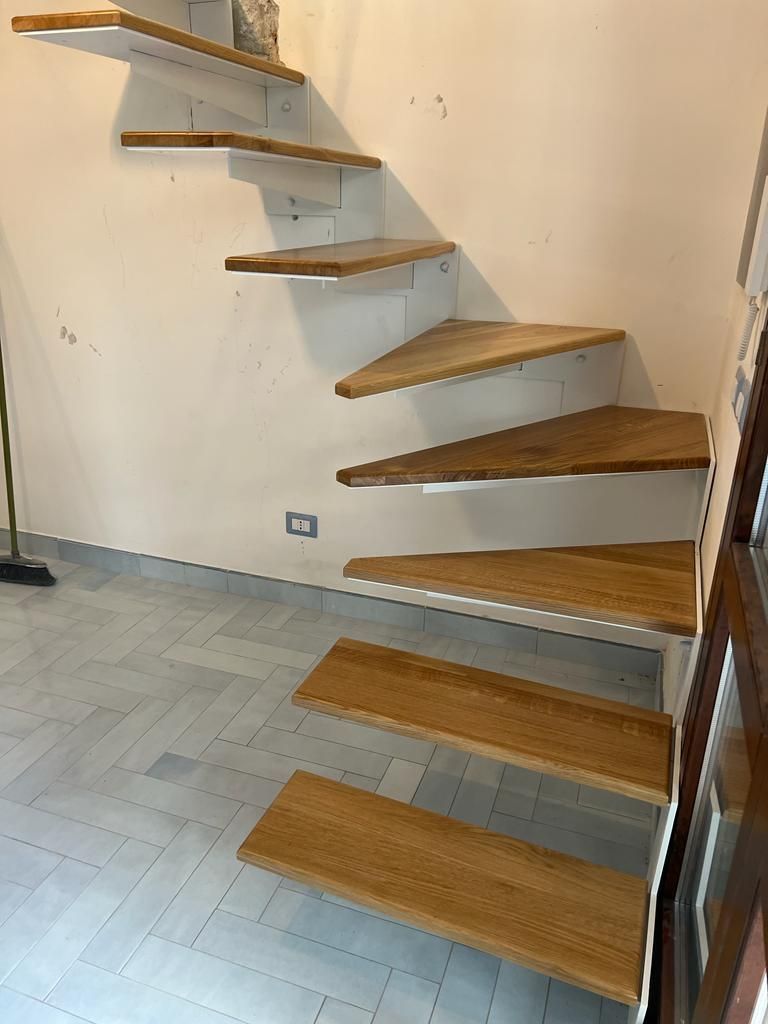 Una scala in legno sospesa contro una parete chiara, che si incurva verso l'alto a sinistra sopra un pavimento piastrellato di grigio.