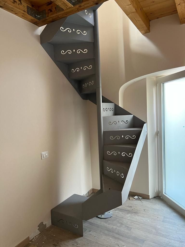 Una scala a chiocciola in metallo grigio con motivi decorativi tagliati al laser sulle alzate, all'interno di una stanza con travi a vista sul soffitto.