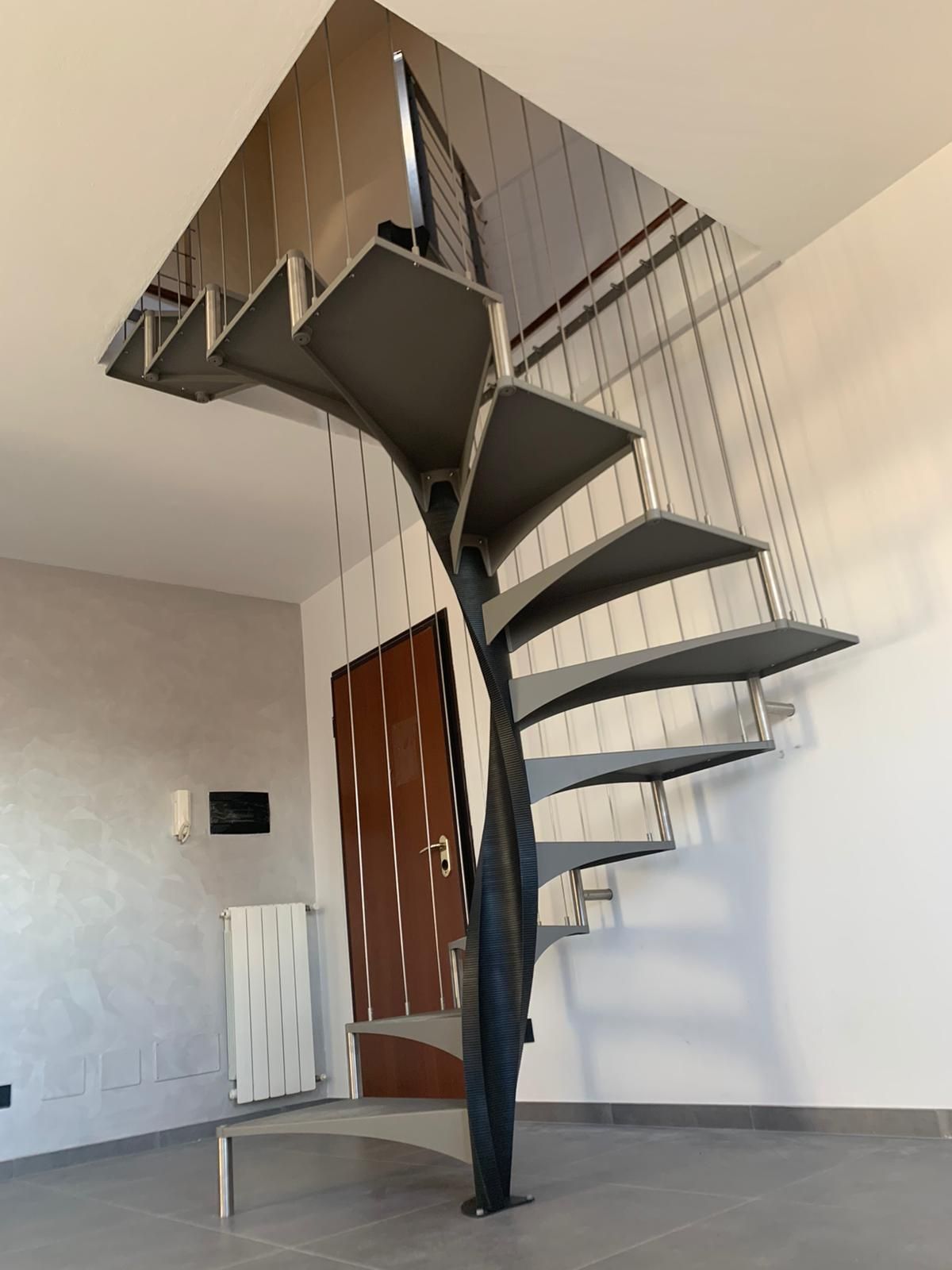 Una moderna scala a chiocciola di colore scuro in una stanza minimalista, con un palo di sostegno centrale e ringhiere a cavo.