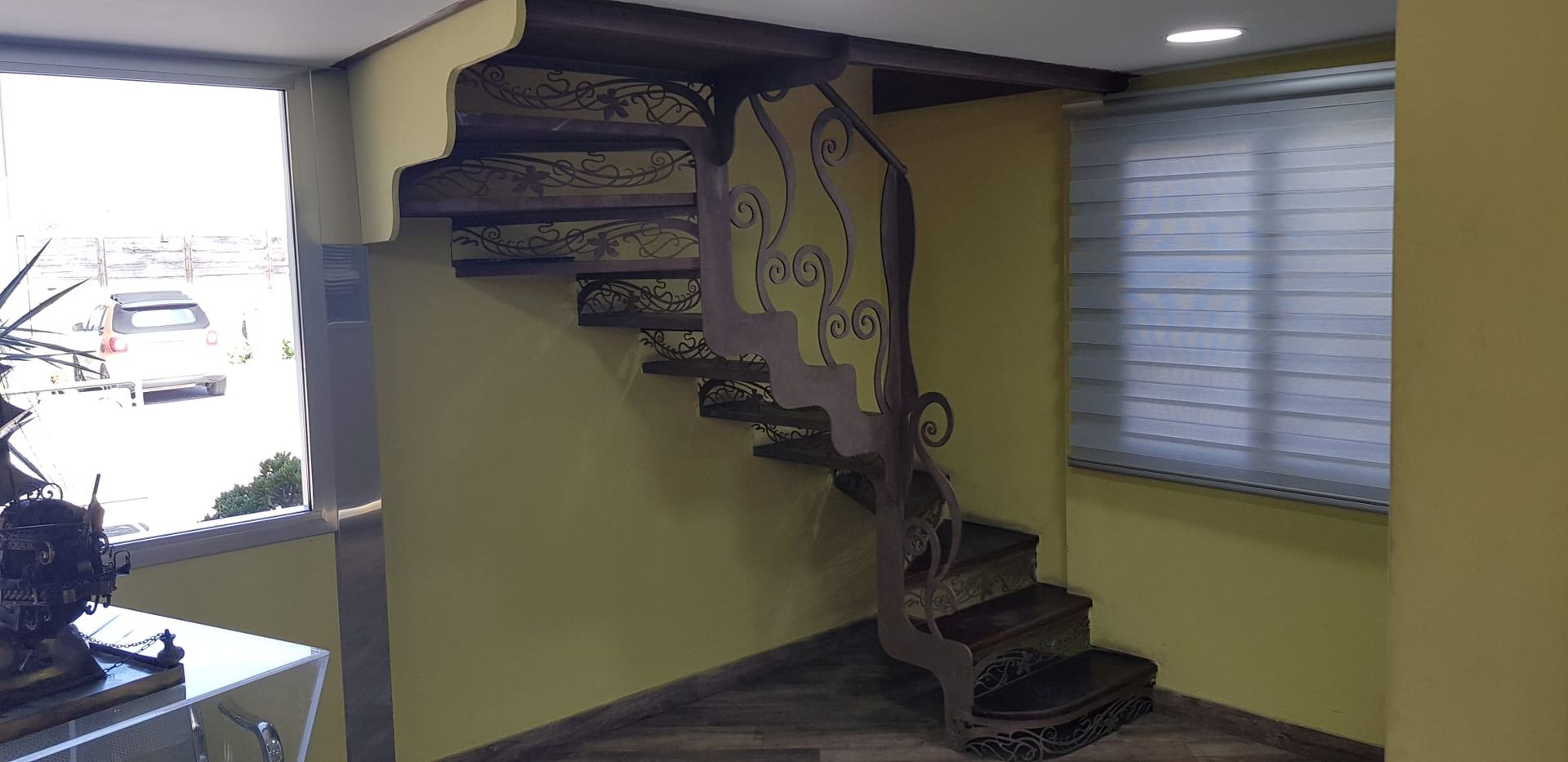 In uno spazio interno, una scala con gradini scuri decorati e una ringhiera metallica decorativa si staglia contro pareti gialle.