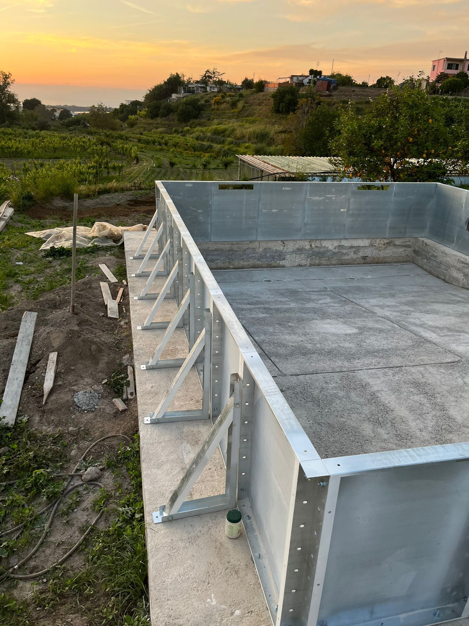 Al tramonto, una struttura metallica per una piscina in costruzione su una base di cemento, con una collina erbosa sullo sfondo.