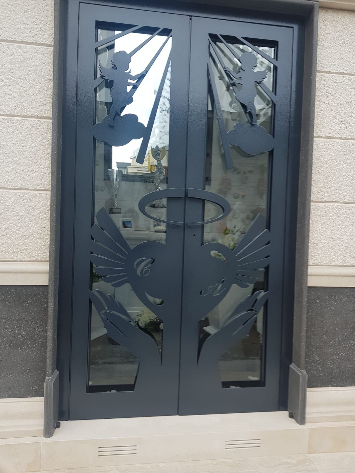 Doppie porte in metallo nero con decorazioni intagliate raffiguranti due angeli su nuvole sopra mani giunte con aureole.