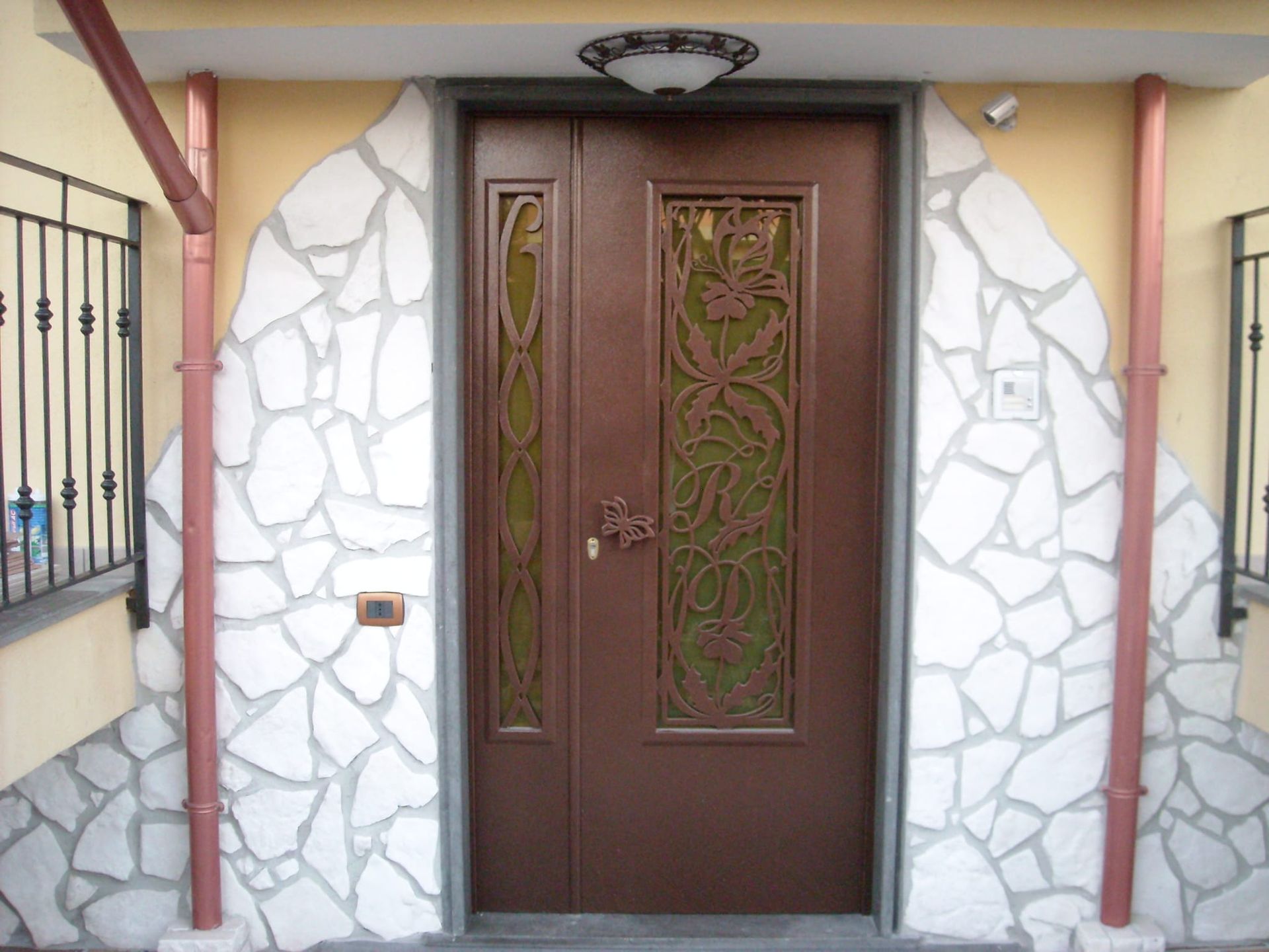 Una porta d'ingresso marrone con due pannelli decorativi in vetro, incorniciata da un rivestimento in pietra bianca e due tubi di scarico in rame.