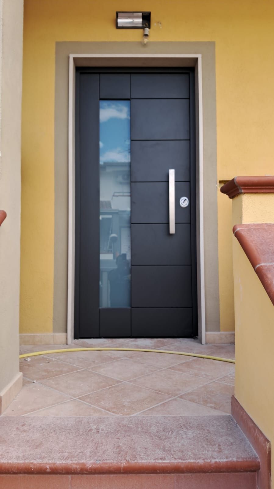 Una moderna porta d'ingresso grigio scuro con pannello verticale in vetro e maniglia argentata, incastonata in un muro giallo sopra una scalinata in pietra.