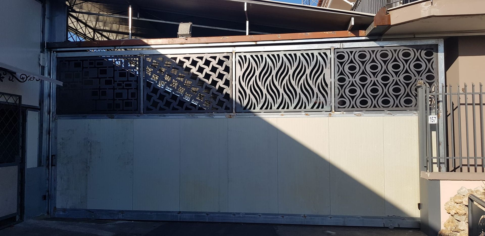 Un ampio cancello carrabile in metallo beige con quattro distinti motivi geometrici tagliati al laser nella parte superiore.