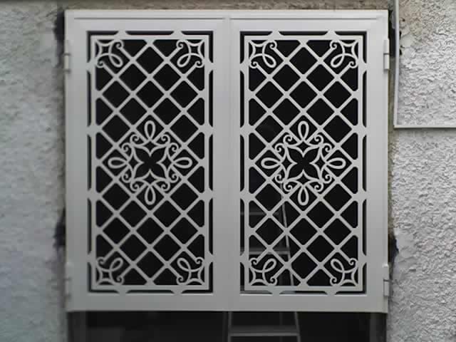 Un cancelletto di sicurezza decorativo in metallo bianco con motivo a griglia e decorazioni floreali, installato sopra una finestra esterna.