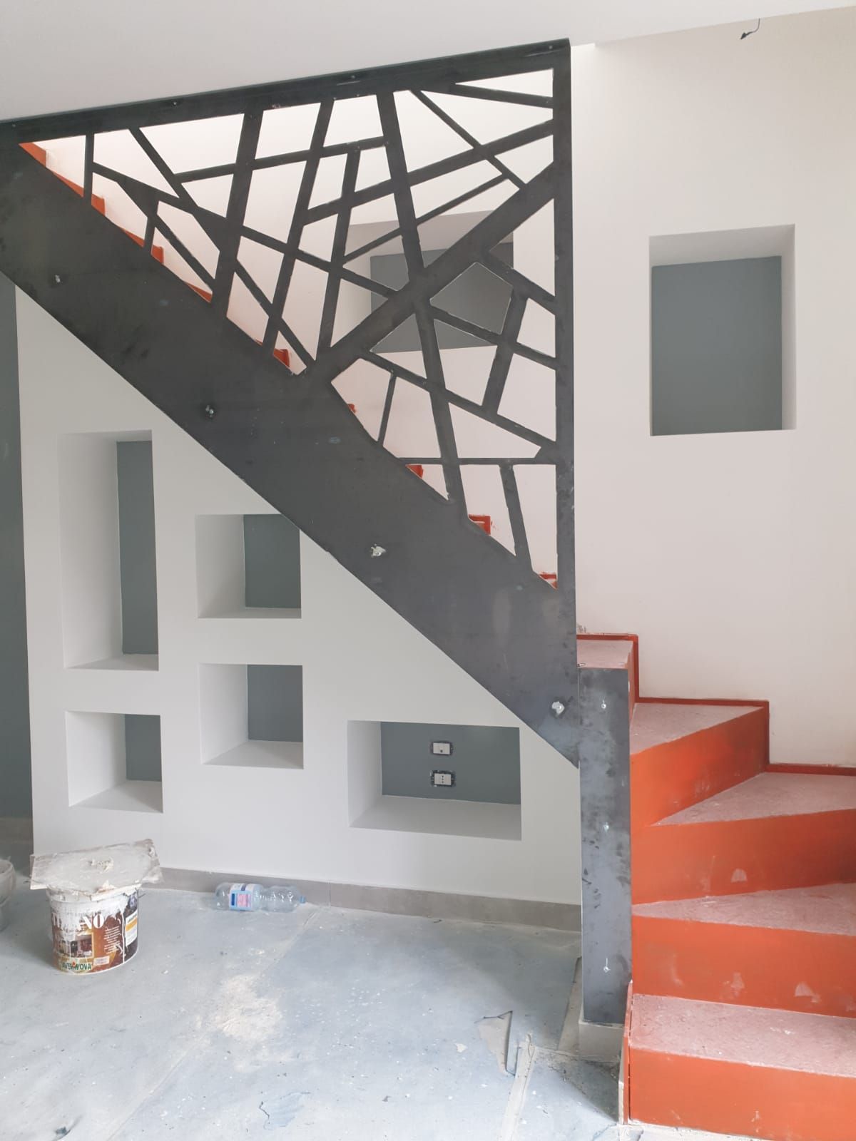 Una scala con gradini arancioni, una ringhiera geometrica in metallo nero e nicchie a muro bianche integrate, in una stanza in fase di ristrutturazione.