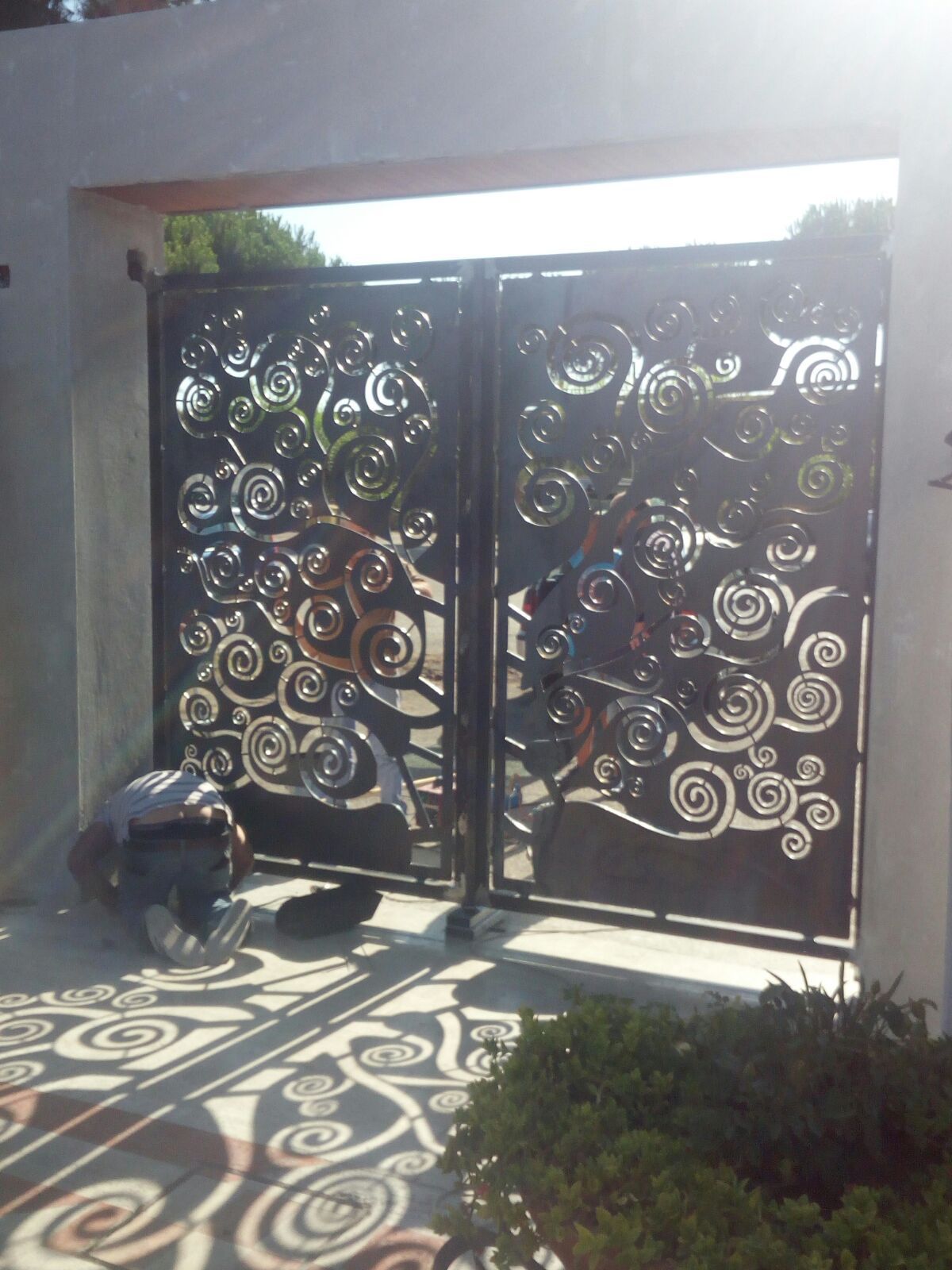 Un cancello in metallo scuro e ornato, con intricati motivi a volute, proietta ombre dettagliate sul terreno in un ingresso illuminato dal sole.