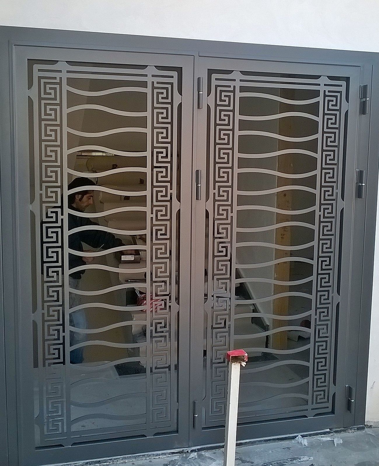 Doppie porte grigie con cornice geometrica a greca e barre metalliche orizzontali ondulate, installate in un ambiente residenziale.