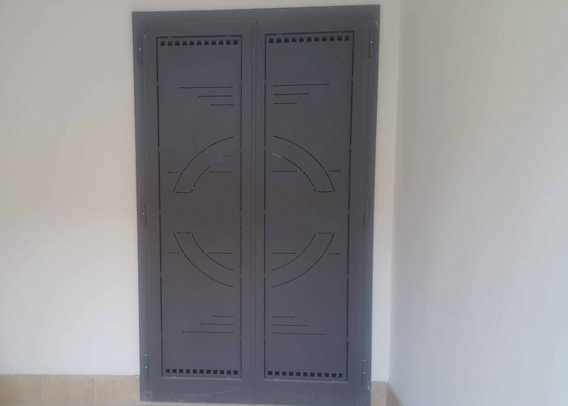 Una porta metallica grigia a doppio pannello con un motivo geometrico intagliato di archi e linee orizzontali, montata su una parete bianca.