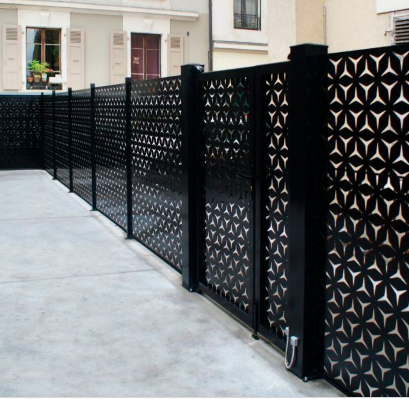 In un contesto urbano, una recinzione metallica nera con un motivo geometrico a intagli ripetuti corre lungo un vialetto di cemento.