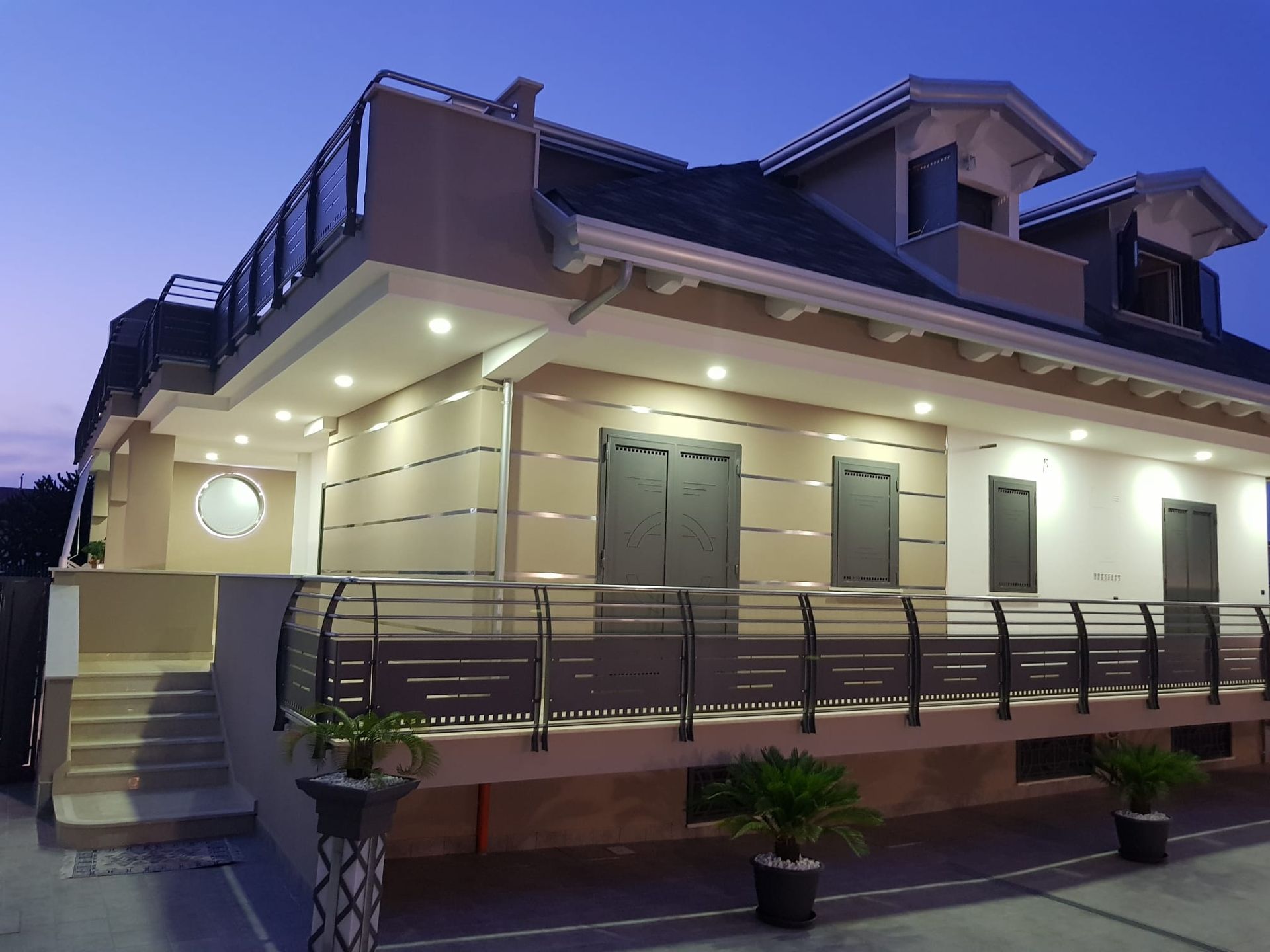 Una casa moderna a due piani al crepuscolo, caratterizzata da esterni beige, una ringhiera metallica sul terrazzo e una calda illuminazione a incasso.