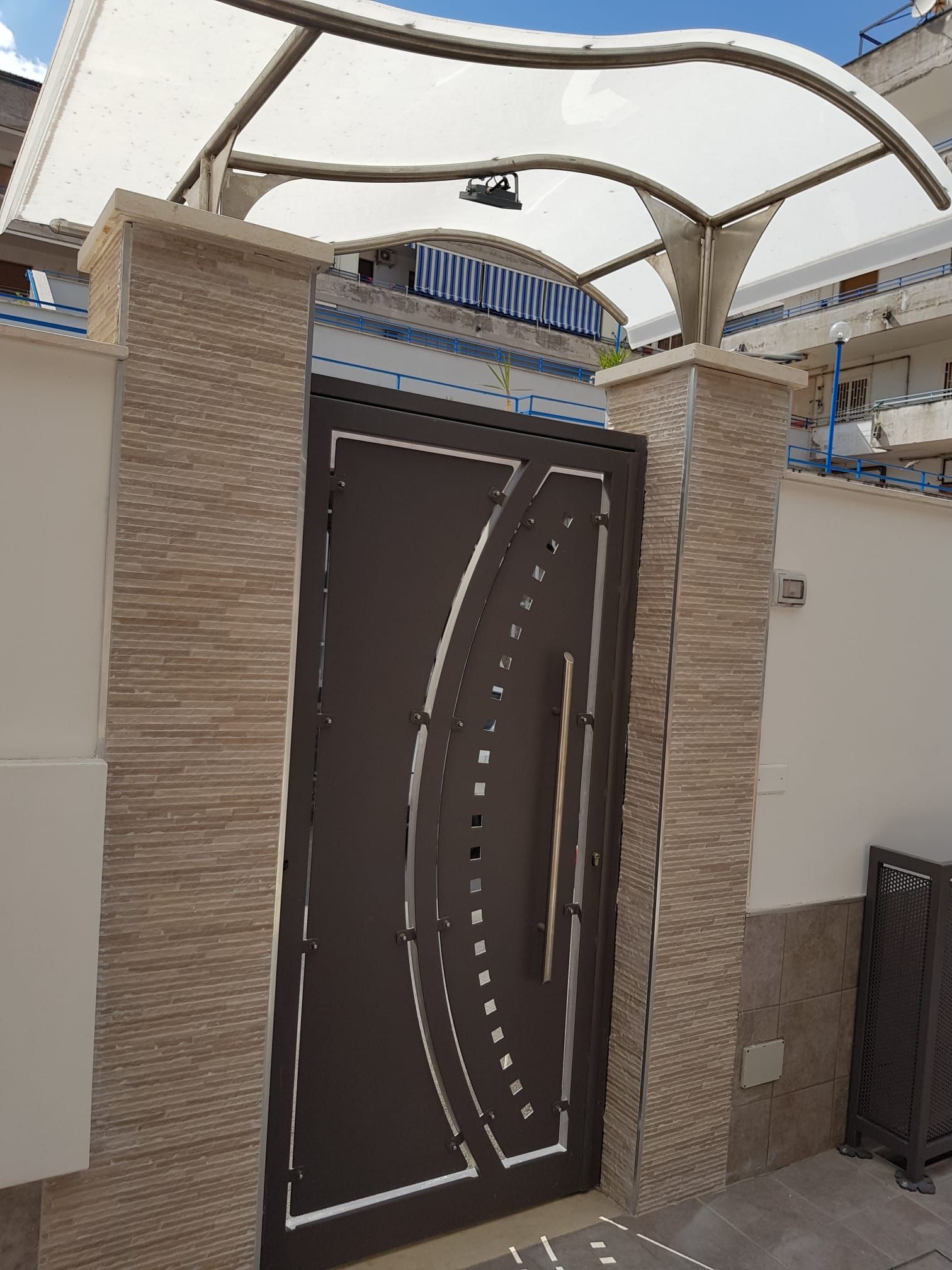 Una moderna porta pedonale in metallo marrone scuro, incastonata tra pilastri con finitura effetto pietra e sormontata da una tettoia curva in tessuto bianco.