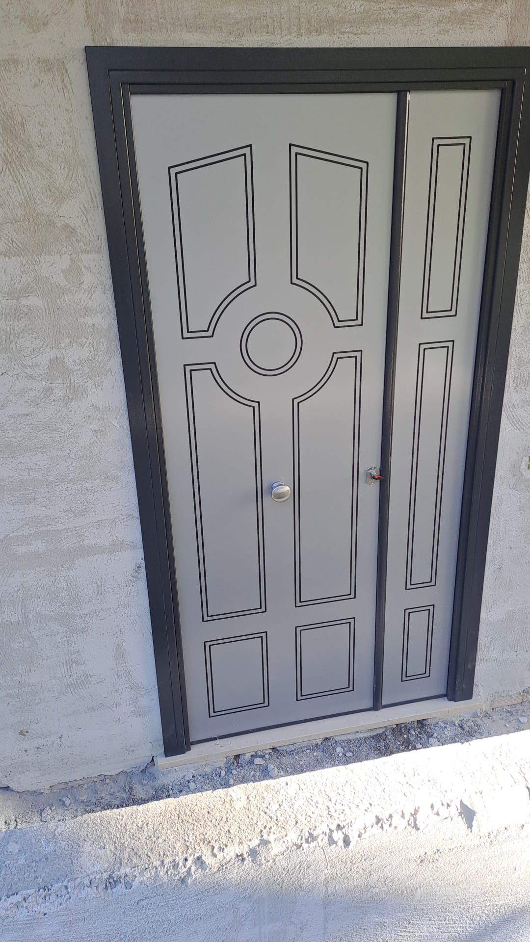 Porta d'ingresso in metallo con pannelli decorativi incastonati in un muro di cemento, illuminata dalla luce del sole.