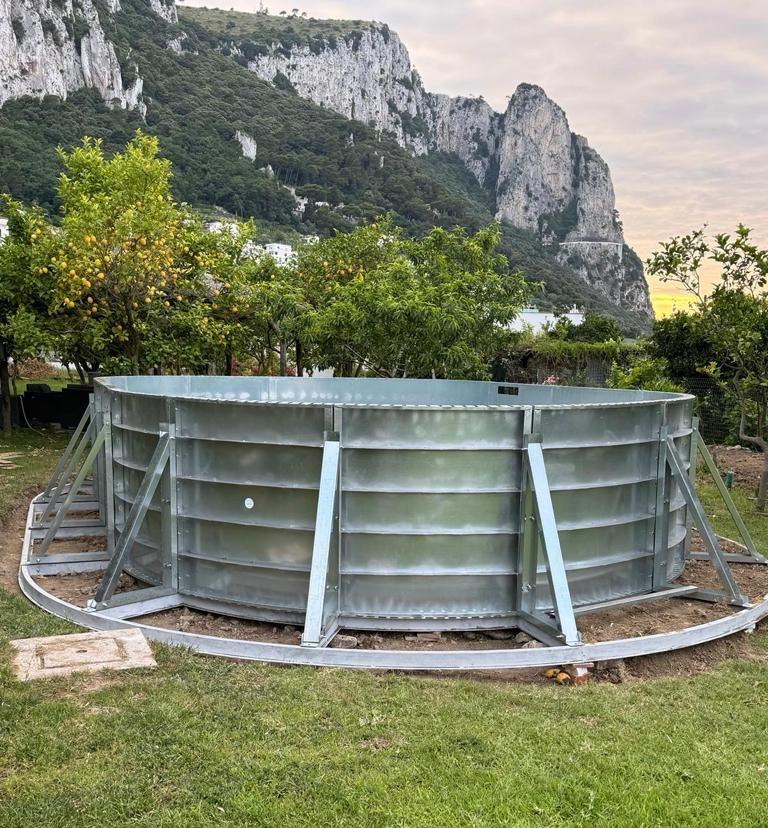 Una base circolare in acciaio zincato per una cisterna d'acqua, con supporti strutturali, immersa in un paesaggio erboso ai piedi di una montagna rocciosa.