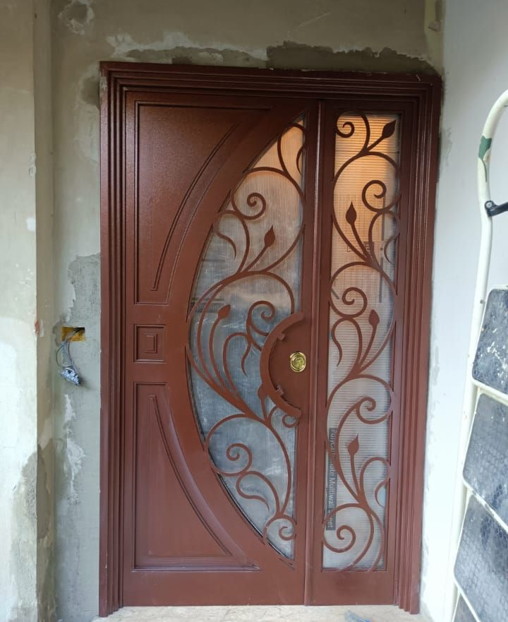 Una porta d'ingresso marrone con un inserto semicircolare in vetro decorato con elaborate volute in metallo, inserita in un vano porta non rifinito.