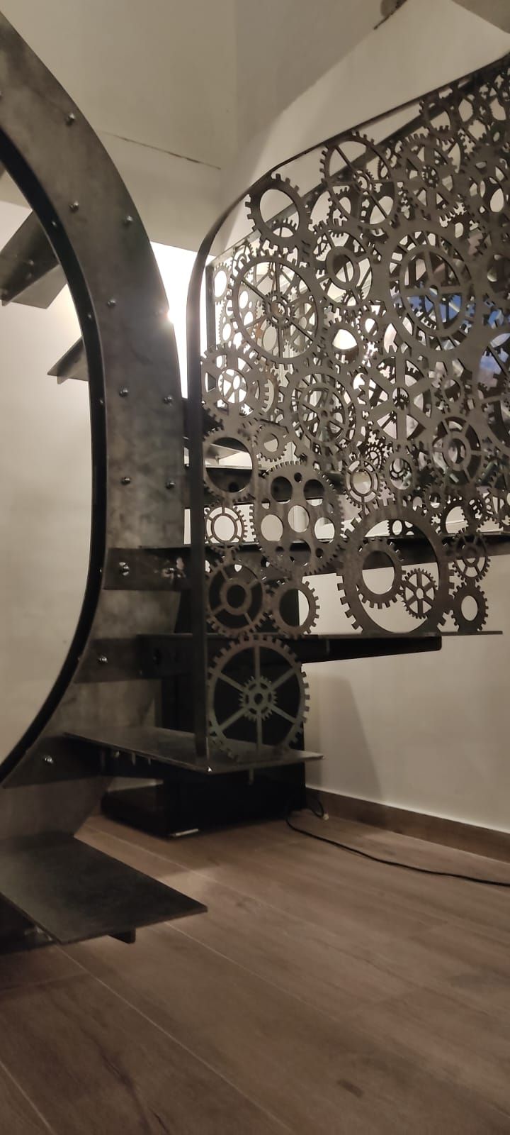 Una scala a chiocciola con ringhiera in stile steampunk composta da ingranaggi metallici ad incastro, in una stanza con pavimento in legno chiaro.