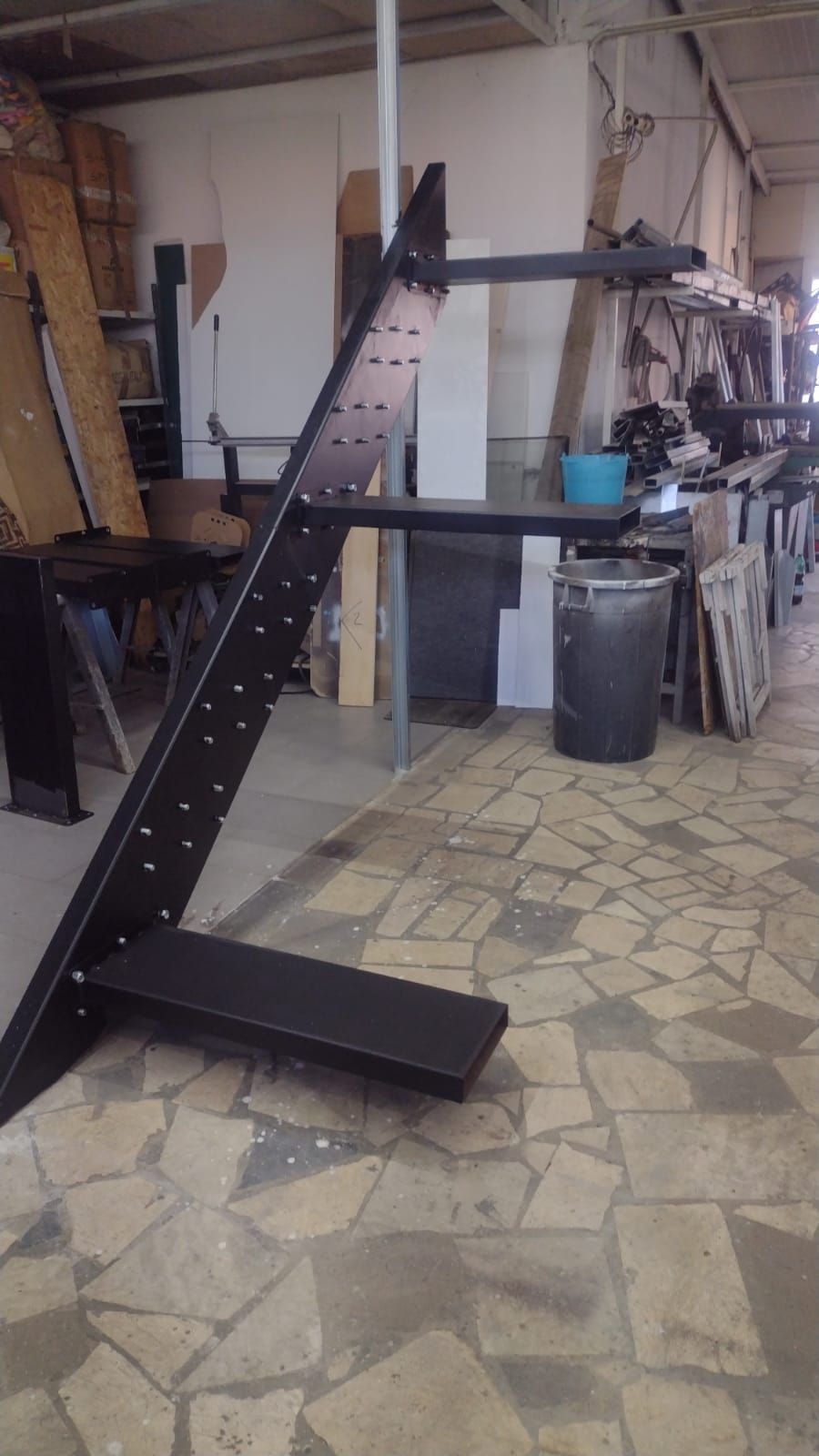 Una struttura portante in metallo nero per una scala, con gradini annessi, posizionata verticalmente in un'officina con pavimento in pietra.