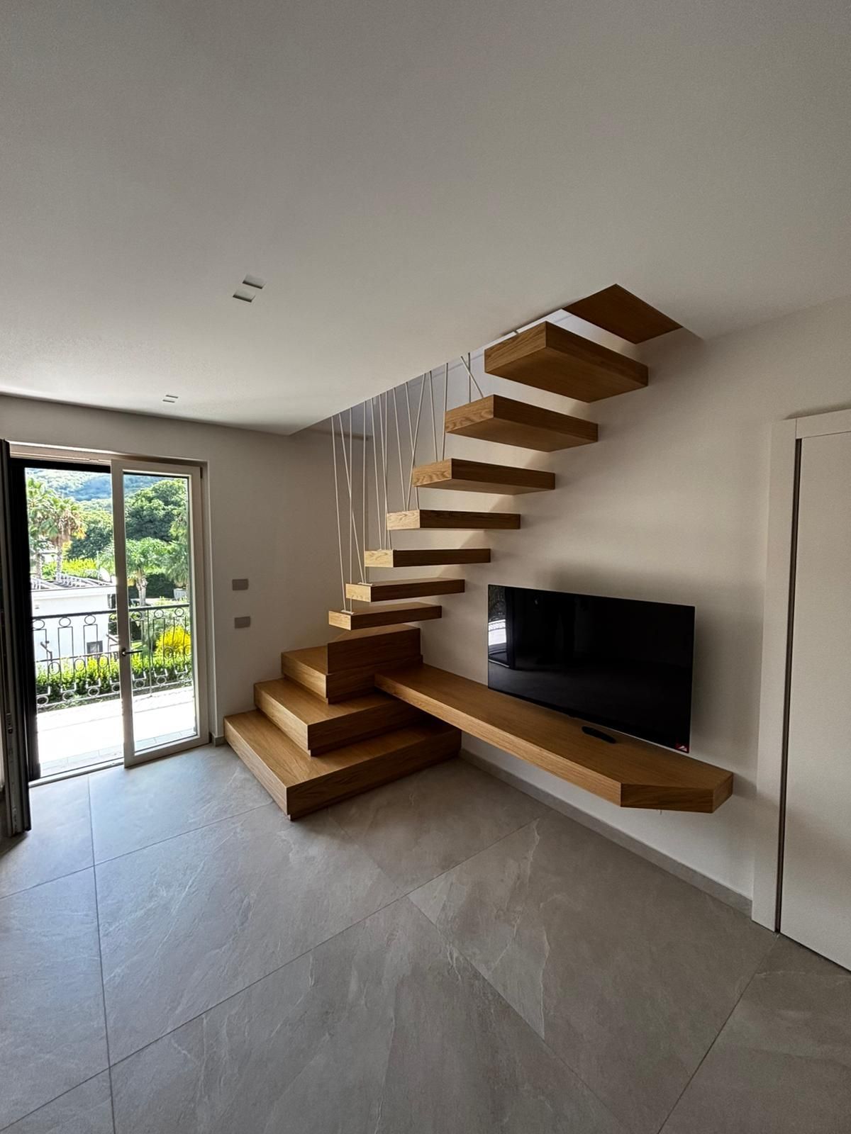 Un soggiorno moderno presenta pavimenti in piastrelle di colore chiaro, un televisore su una mensola di legno sospesa e una scala in legno sospesa.