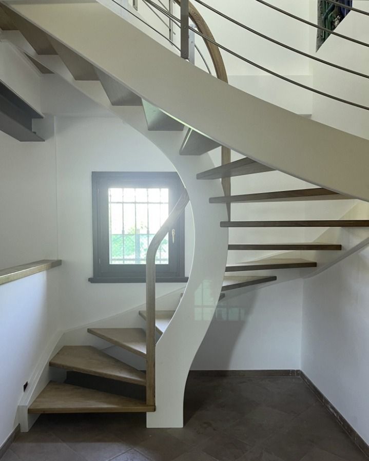 Una moderna scala curva bianca con gradini in legno e una sottile ringhiera in metallo, situata in una stanza con una finestra con cornice scura.