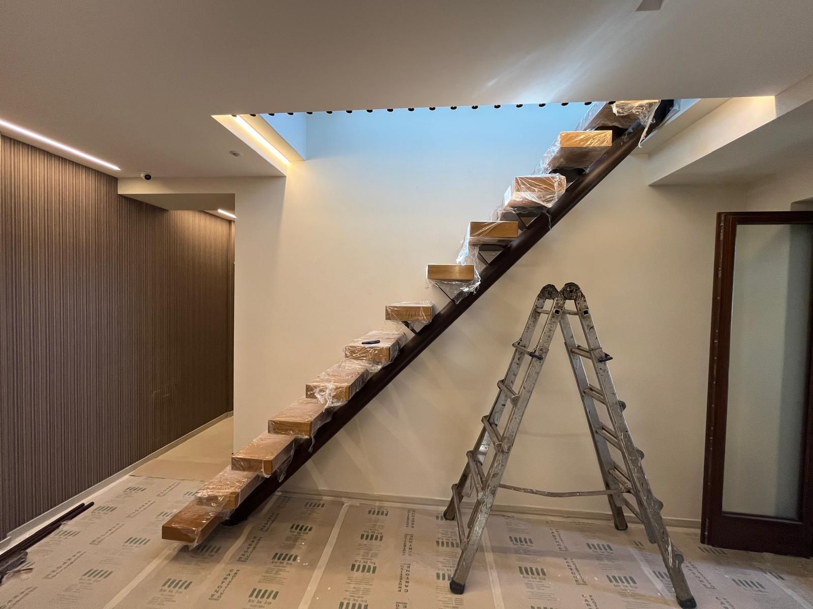 Una scala in costruzione con gradini in legno fissati a una struttura metallica scura, con una scala a pioli in metallo nelle vicinanze.