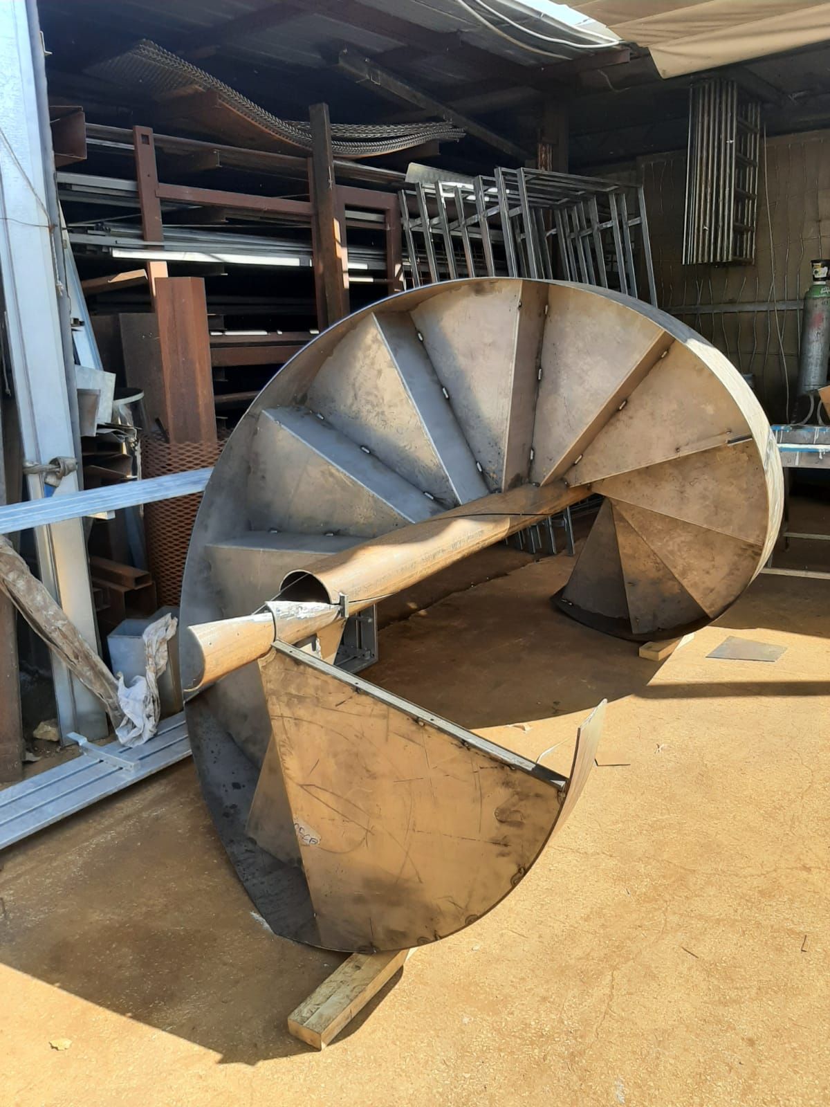 Una scala a chiocciola in metallo giace su un fianco in un'area di stoccaggio esterna piena di materiali metallici.