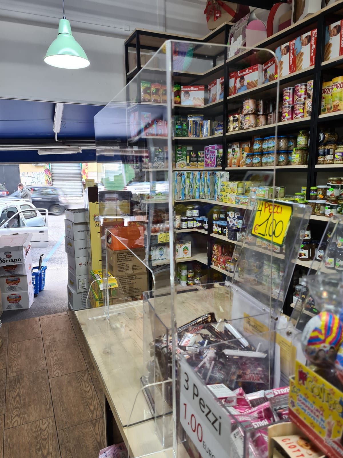 Il bancone di un negozio di alimentari con una barriera in plexiglass trasparente, scaffali pieni di vari prodotti e un cartellino del prezzo.