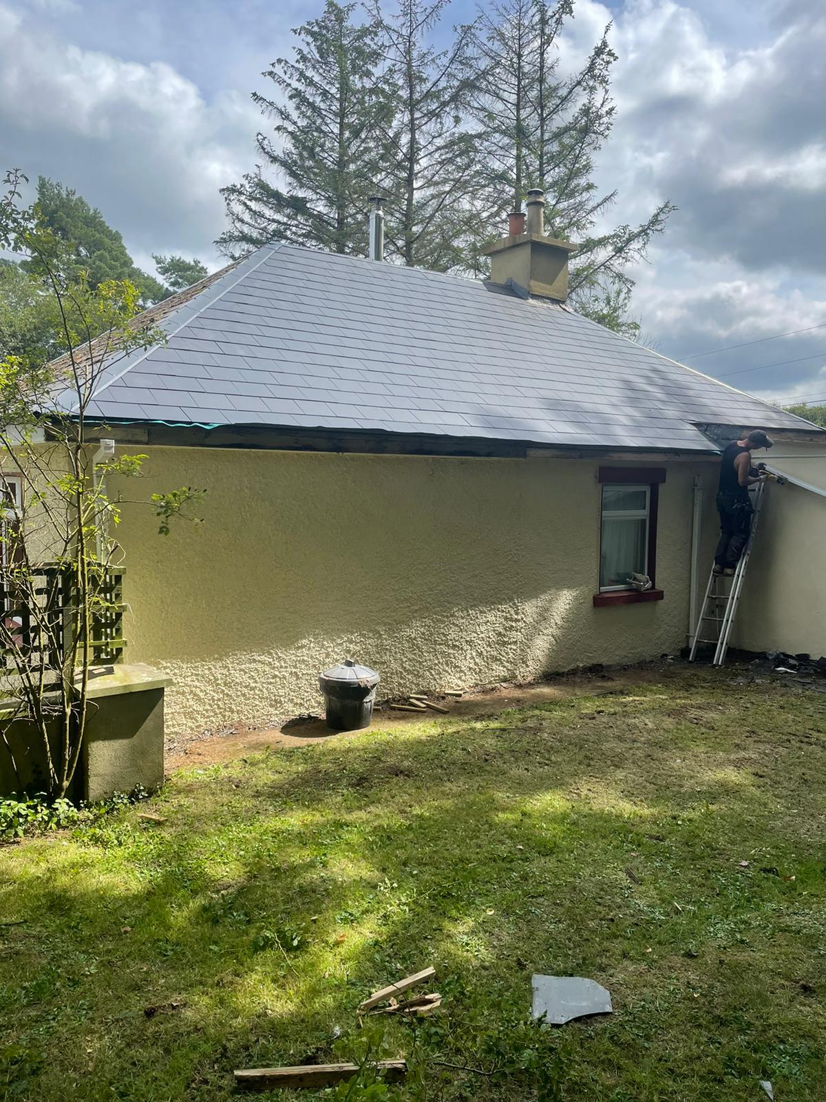 A man is standing on a ladder in front of a house with a roof that is being painted.