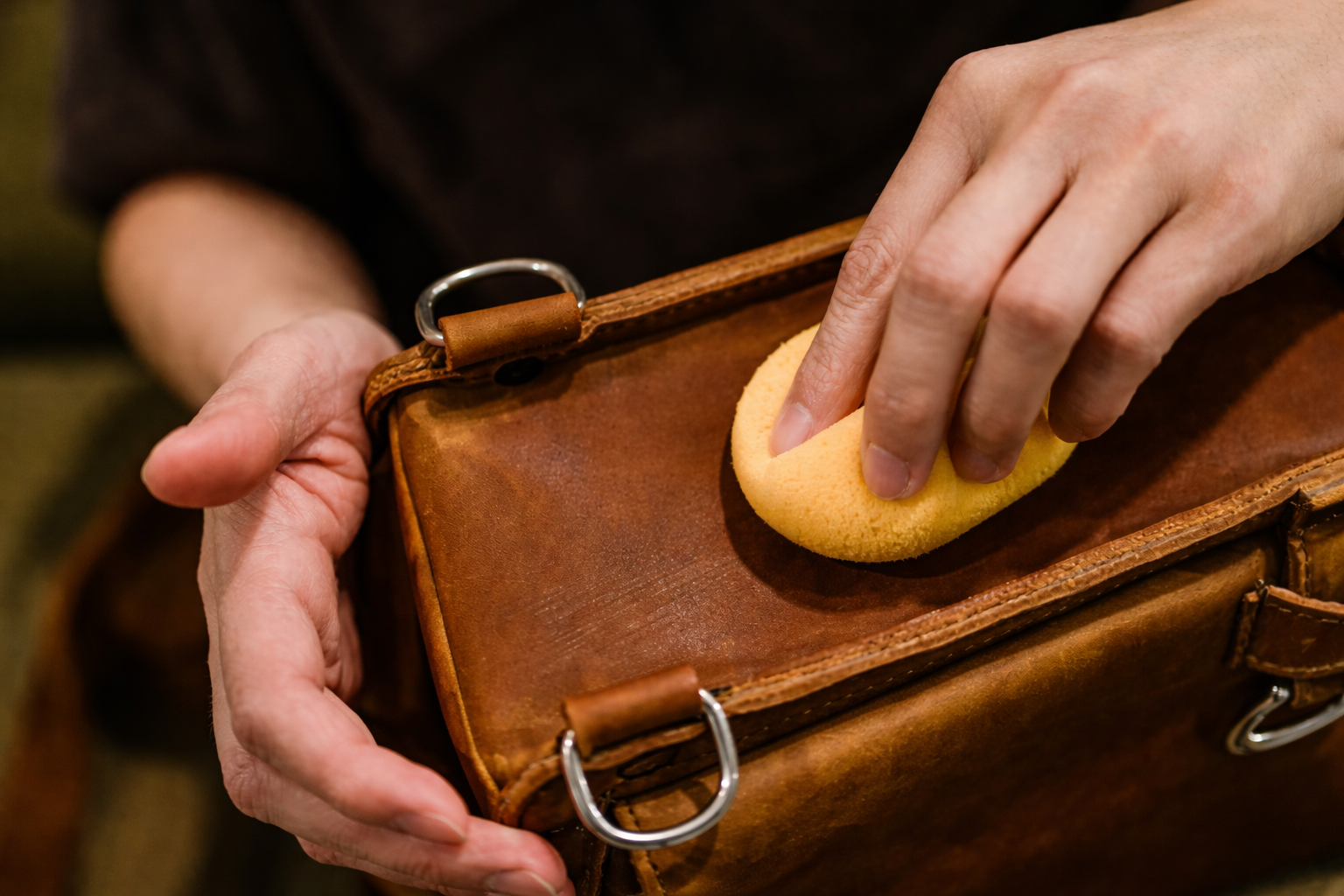 Hands use a yellow sponge to apply conditioner to the surface of a brown leather bag.