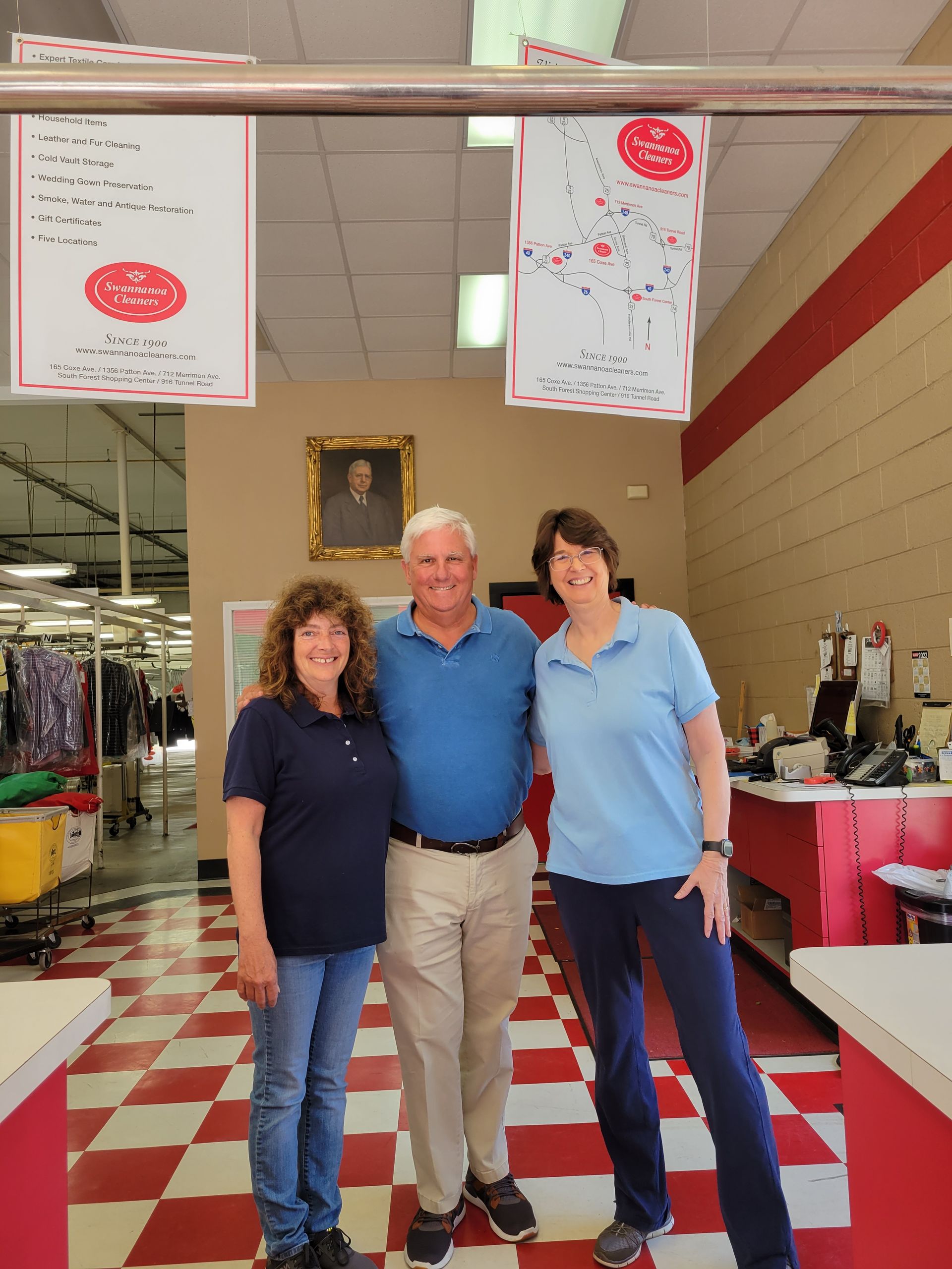 Three people stand smiling in a dry cleaners with red-and-white tiled floors, between two hanging signs.