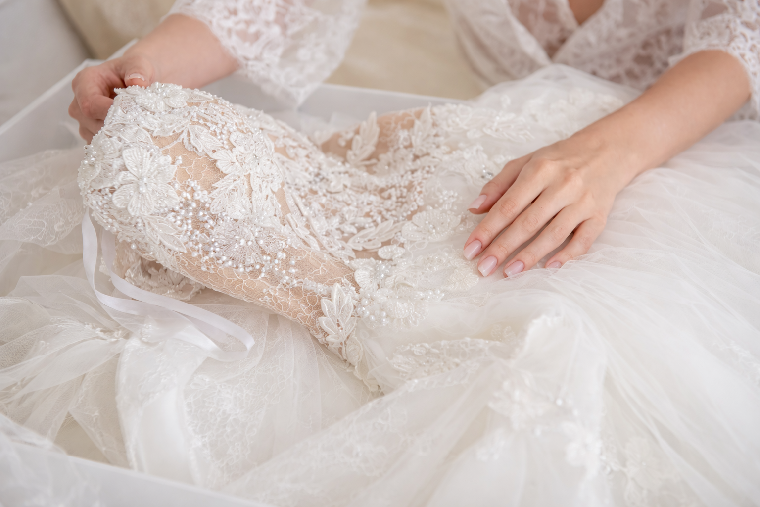 Hands gently touch an intricate white lace wedding dress as it is laid out inside a box.