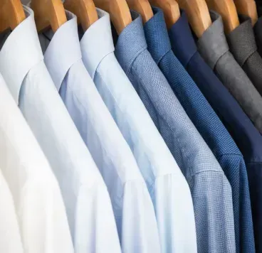 A row of men's button-down shirts in shades of white, light blue, and dark blue hanging on wooden hangers.