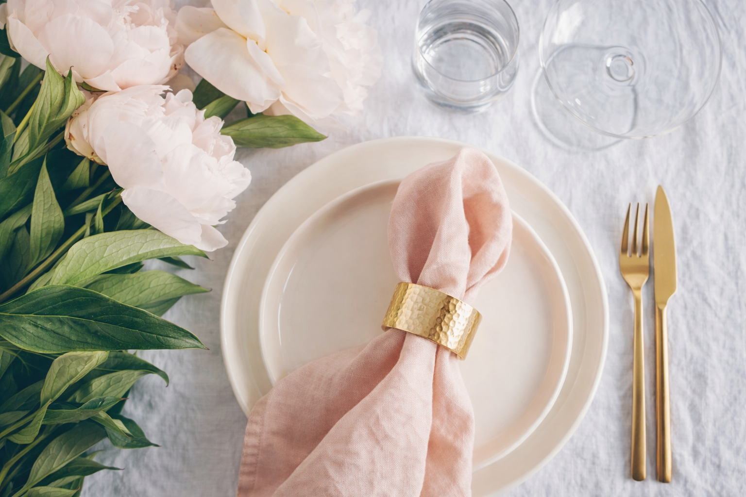 A light pink napkin in a gold napkin ring sits on a cream plate next to a gold fork, knife, and a bunch of pink peonies.