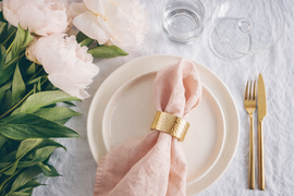 A light pink napkin in a gold napkin ring sits on a cream plate next to a gold fork, knife, and a bunch of pink peonies.