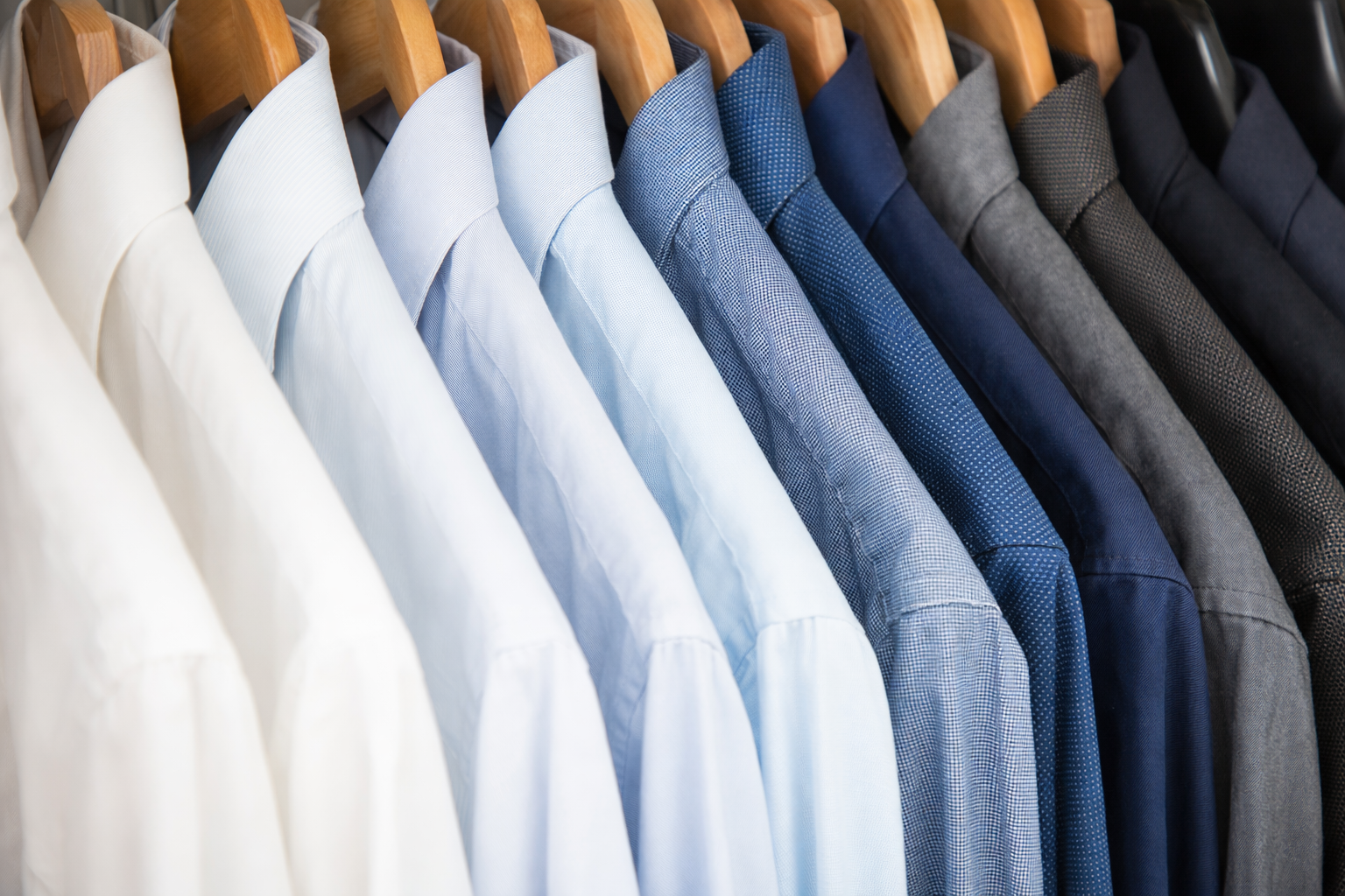 A row of men's button-down shirts in shades of white, light blue, and dark blue hanging on wooden hangers.