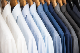 A row of men's button-down shirts in shades of white, light blue, and dark blue hanging on wooden hangers.
