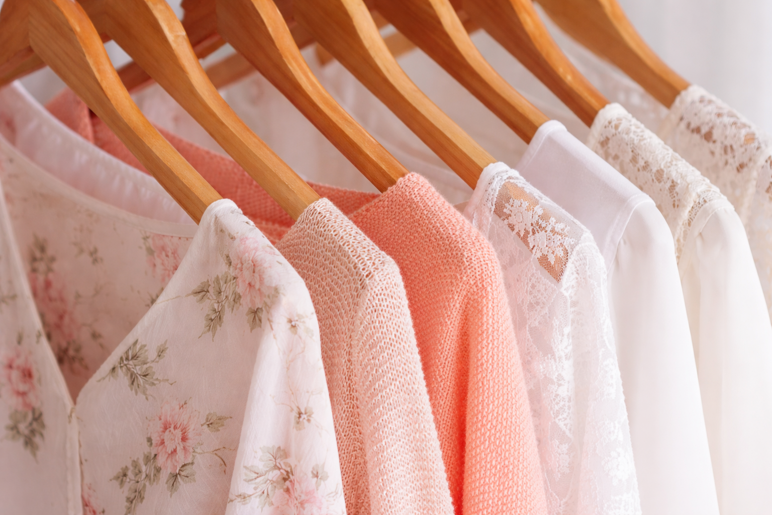Several light-colored shirts and cardigans with floral, textured, and lace patterns hanging on wooden hangers.
