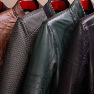 Close-up of four leather jackets in tan, textured grey, green, and black, hanging on red hangers.