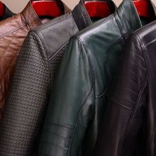 Close-up of four leather jackets in tan, textured grey, green, and black, hanging on red hangers.