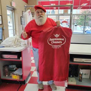 Santa Claus in red clothing stands in a shop, holding a branded garment bag from Swannanoa Cleaners and giving a thumbs-up.