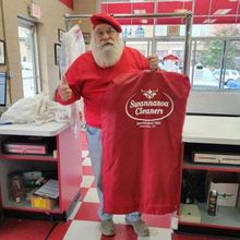 Santa Claus in red clothing stands in a shop, holding a branded garment bag from Swannanoa Cleaners and giving a thumbs-up.